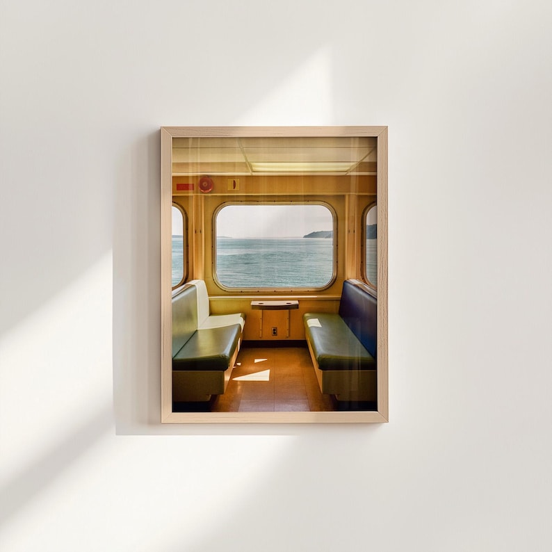 May include: A framed wall art print depicting the interior of a ferry cabin with yellow walls, a central window overlooking the sea, and two rows of bench seating in green and blue, all encased in a light-colored wooden frame.