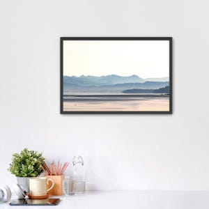 May include: A framed landscape print featuring a serene lake reflecting soft light, with layered blue mountain silhouettes in the distance under a bright sky. The artwork is enclosed in a thin black frame, showcasing a calm, atmospheric view of water and distant peaks.