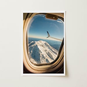 May include: A photograph taken from an airplane window, showcasing a snow-covered mountain range under a clear blue sky. The wing of the plane is visible, with a green and blue design on the wingtip.