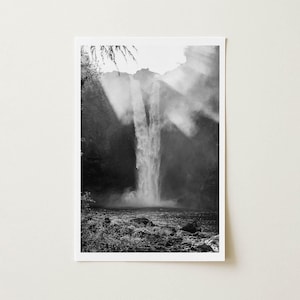 May include: A black and white vertical photograph depicting a tall waterfall cascading down a dark, rocky cliff face. Mist rises from the base where the water meets a pool, while bright light streams from above, illuminating the falling water.