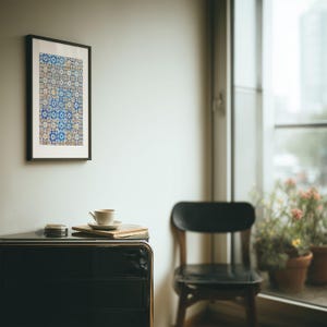 May include: A framed artwork with a blue and white geometric pattern hangs on a white wall. A black cabinet with a coffee cup, saucer, and book sits below. A black chair and potted plants are visible.