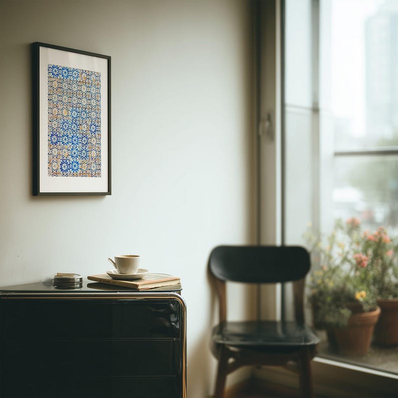May include: A framed artwork with a blue and white geometric pattern hangs on a white wall. A black cabinet with a coffee cup, saucer, and book sits below. A black chair and potted plants are visible.