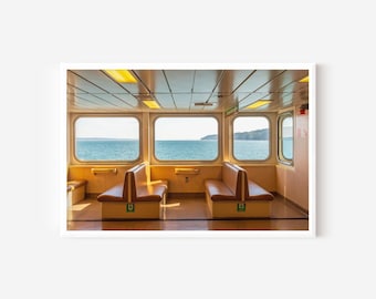May include: The interior of a ferry cabin features two rows of tan vinyl bench seating facing each other. Large rectangular windows line the walls, offering a clear view of the blue ocean and distant land under a bright, natural daylight sky.