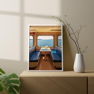 May include: A framed wall art print featuring the interior of a ferry cabin with two blue bench seats facing each other across a small table. Large windows reveal a view of the blue ocean and distant shoreline under a clear sky.