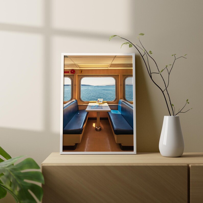 May include: A framed wall art print featuring the interior of a ferry cabin with two blue bench seats facing each other across a small table. Large windows reveal a view of the blue ocean and distant shoreline under a clear sky.