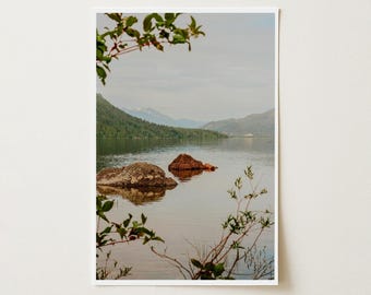 May include: A landscape photograph of a calm lake with two prominent rocks emerging from the water. In the background, green forested hills rise toward distant mountains under a soft, overcast sky. Green leafy branches frame the foreground.