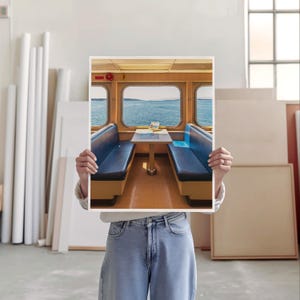 May include: A rectangular art print featuring a photograph of a ferry interior with two blue bench seats facing each other across a central table. Large windows reveal a view of the blue ocean and distant horizon under a bright sky.
