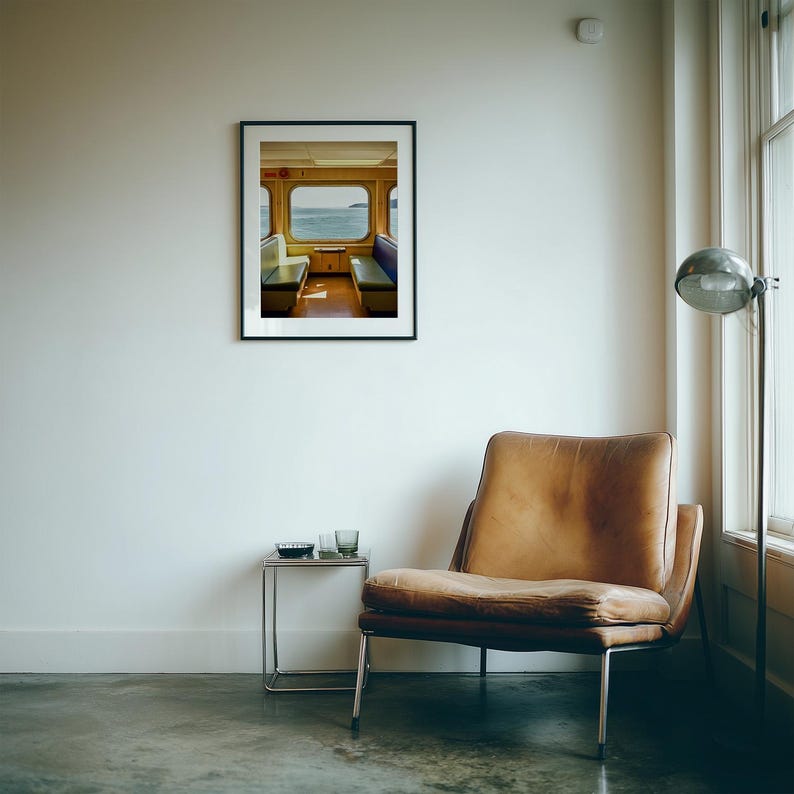 May include: A framed wall art print depicting the interior of a train carriage with two rows of seats facing a window overlooking the sea. Below the print sits a brown leather armchair and a small metal side table with two glasses.