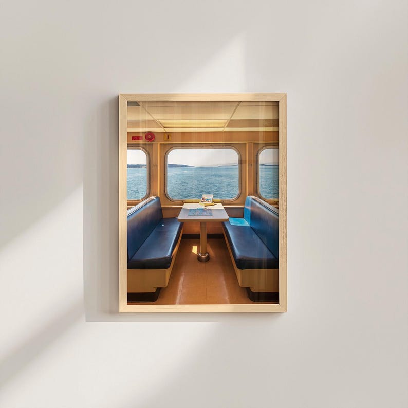 May include: A framed wall art print depicting the interior of a ferry cabin with two rows of dark blue bench seating facing each other across a central table. Large windows reveal a view of the open sea and distant horizon under a clear sky.