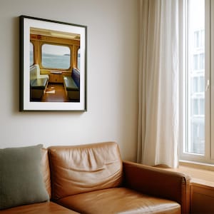 May include: A framed wall art print depicting the interior of a ferry or train cabin with yellow walls, two rows of bench seating in blue and white, and large windows looking out onto a body of water under a bright, overcast sky.