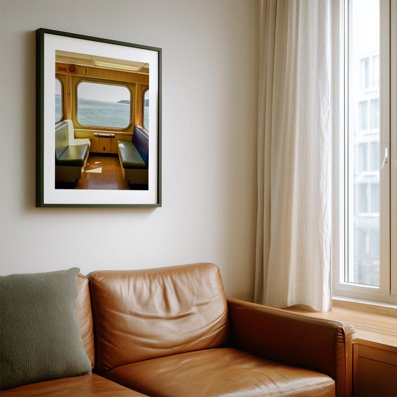 May include: A framed wall art print depicting the interior of a ferry or train cabin with yellow walls, two rows of bench seating in blue and white, and large windows looking out onto a body of water under a bright, overcast sky.