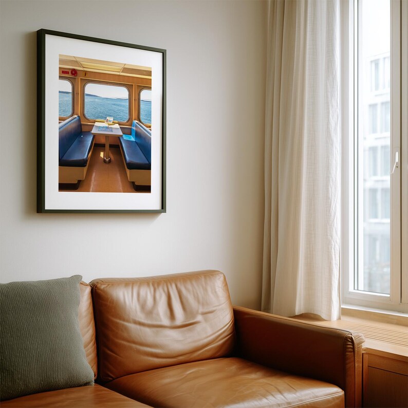 May include: A framed wall art print depicting the interior of a ferry cabin with two blue bench seats facing each other across a small table, positioned in front of large windows that reveal a view of the open sea and distant shoreline.