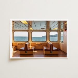 May include: A photograph of a ferry interior featuring two rows of tan vinyl bench seating facing each other. Large rectangular windows reveal a view of the ocean and distant shoreline. The ceiling has recessed rectangular lighting.