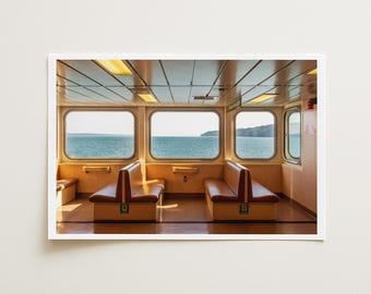 May include: A photograph of a ferry interior featuring two rows of tan vinyl bench seating facing each other. Large rectangular windows reveal a view of the ocean and distant shoreline. The ceiling has recessed rectangular lighting.
