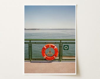 May include: A bright orange life ring with a peace sign center is mounted on a green metal railing overlooking a calm body of water. The life ring features the text M/V SUQUAMISH SEATTLE printed in black lettering along its circular frame.