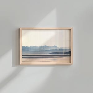 May include: A rectangular landscape print in a light-toned wooden frame featuring a serene view of a calm lake reflecting the soft light of dawn or dusk, with layered blue mountain silhouettes in the distance under a pale, hazy sky.