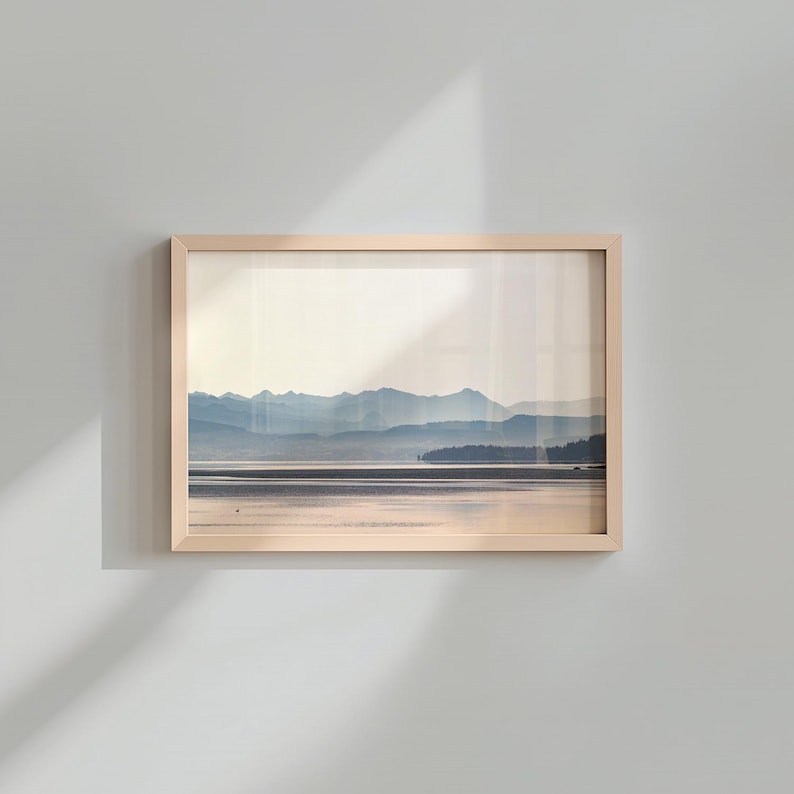 May include: A rectangular landscape print in a light-toned wooden frame featuring a serene view of a calm lake reflecting the soft light of dawn or dusk, with layered blue mountain silhouettes in the distance under a pale, hazy sky.