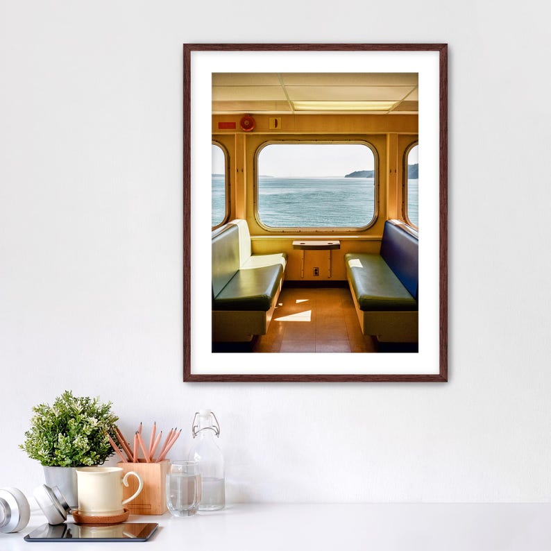 May include: A framed wall art print featuring the interior of a ferry cabin with two rows of green bench seating facing a large rectangular window that overlooks a calm body of water under a bright sky, set in a dark brown wooden frame.
