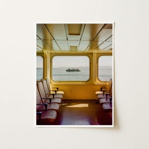 May include: A view from inside a ferry cabin featuring rows of brown upholstered seats facing large rectangular windows. Through the center window, a white ferry boat is visible on the water under a bright sky, with a light-colored ceiling.