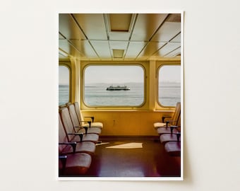 May include: A view from inside a ferry cabin featuring rows of brown upholstered seats facing large rectangular windows. Through the center window, a white ferry boat is visible on the water under a bright sky, with a light-colored ceiling.