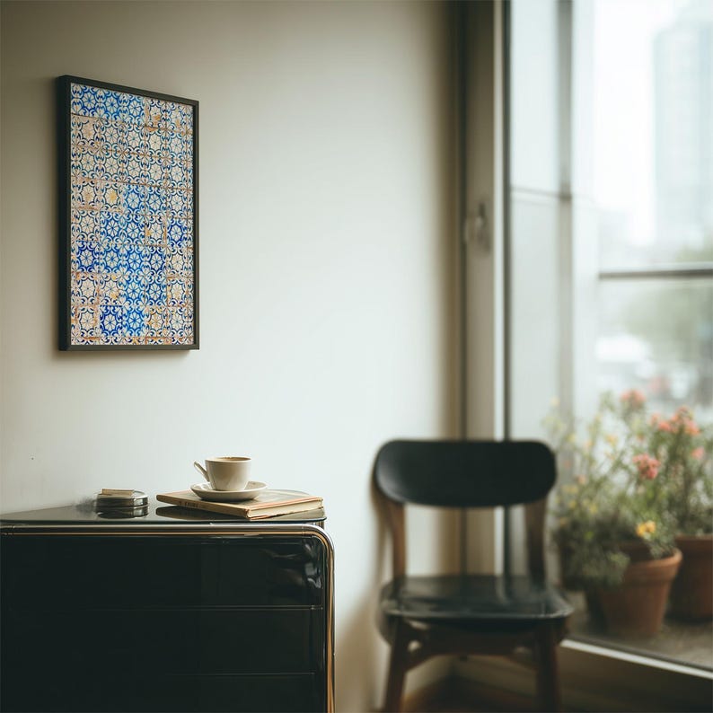 May include: A framed artwork with a detailed, colorful tile pattern hangs on a white wall. A black cabinet with a cup and saucer, coasters, and a book sits below. A black chair and window with plants are also visible.