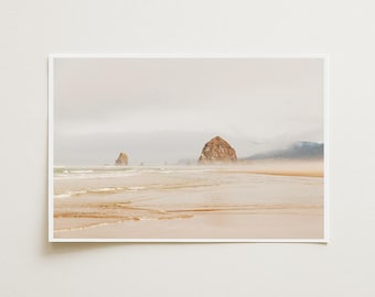 May include: A landscape photograph of a sandy beach with wet, reflective sand in the foreground. Two large, jagged rock formations rise from the ocean in the distance under a thick, overcast, and foggy sky with muted beige and grey tones.