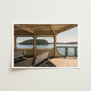 May include: A view from the interior deck of a ferry featuring beige metal benches, a cream-colored ceiling structure, and large rectangular windows looking out onto a calm body of water with a small, tree-covered island under a clear sky.