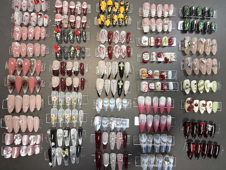 Puede incluir: Una muestra de 20 filas de u&ntilde;as postizas con varios dise&ntilde;os y colores. Las u&ntilde;as est&aacute;n dispuestas en filas sobre un fondo blanco. Las u&ntilde;as son de diferentes formas y tama&ntilde;os, y algunas tienen brillo, pedrer&iacute;a u otros adornos.