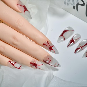 May include: A set of long, pointed artificial nails. The nails are clear with a red abstract design and small silver accents. The nails are displayed on a mannequin hand, with a gold ring with two pearls. The word "HAPPY" is visible in the background.