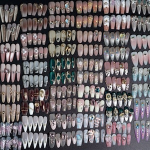 Puede incluir: Una gran colecci&oacute;n de u&ntilde;as postizas coloridas y decoradas. Las u&ntilde;as est&aacute;n dispuestas en filas sobre una superficie negra. Las u&ntilde;as est&aacute;n decoradas con diamantes de imitaci&oacute;n, purpurina y otros adornos.