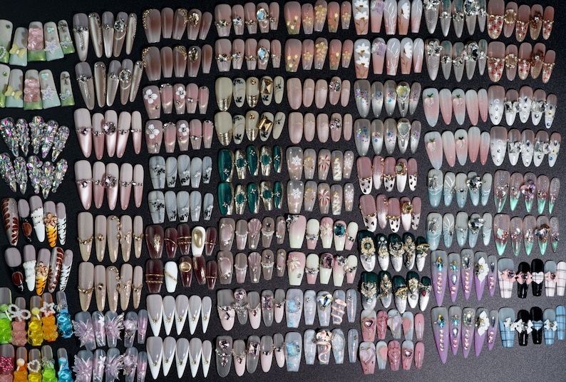 Puede incluir: Una gran colecci&oacute;n de u&ntilde;as postizas coloridas y decoradas. Las u&ntilde;as est&aacute;n dispuestas en filas sobre una superficie negra. Las u&ntilde;as est&aacute;n decoradas con diamantes de imitaci&oacute;n, purpurina y otros adornos.