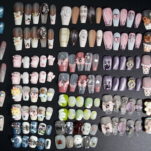 Puede incluir: Una colecci&oacute;n de 20 filas de u&ntilde;as postizas decorativas y coloridas. Cada fila presenta un dise&ntilde;o diferente, que incluye personajes de dibujos animados, corazones, flores y patrones geom&eacute;tricos.