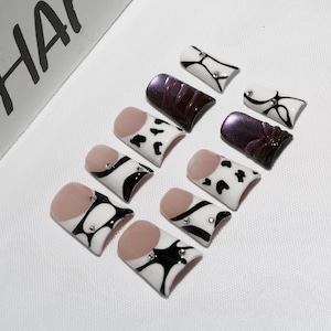 May include: A collection of artificial nails in various designs. Some nails feature a nude base with black and white patterns, while others have a dark purple or white design. Some nails have small, decorative rhinestones. The word "Y HA" is partially visible.