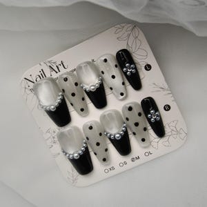 May include: A set of artificial nails with a variety of designs. Some nails are black with pearl accents, others are white with black polka dots, and some have a pearl border. The words "Nail Art" are printed on the card.
