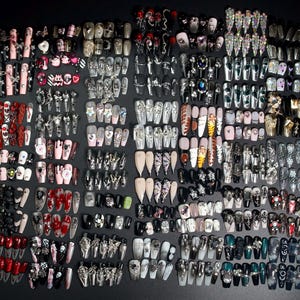 Puede incluir: Una colecci&oacute;n de m&aacute;s de 100 u&ntilde;as postizas coloridas y detalladas con varios dise&ntilde;os, incluyendo calaveras, corazones, estrellas y purpurina. Las u&ntilde;as est&aacute;n dispuestas en filas sobre un fondo negro.