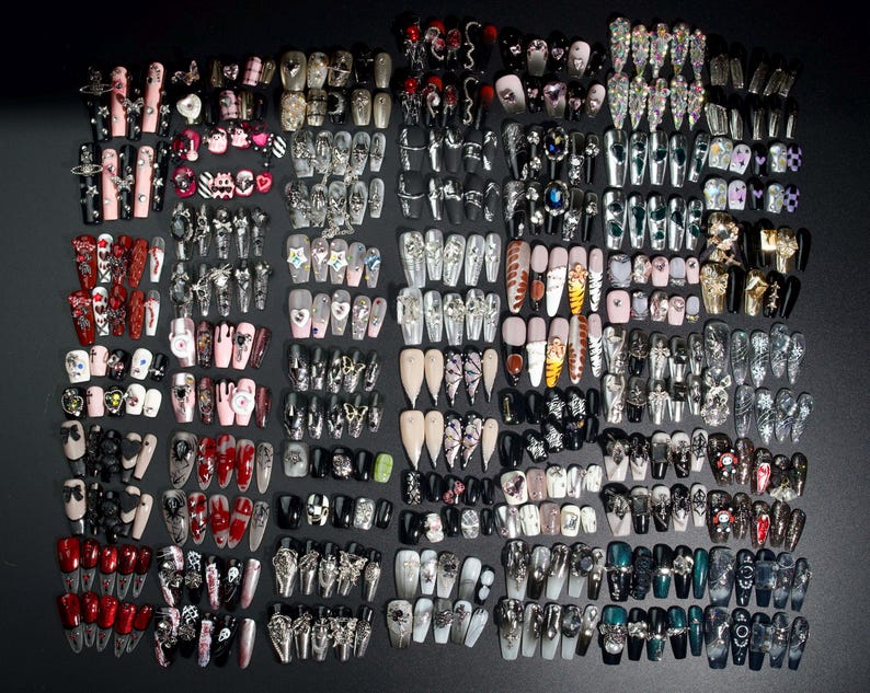 Puede incluir: Una colecci&oacute;n de m&aacute;s de 100 u&ntilde;as postizas coloridas y detalladas con varios dise&ntilde;os, incluyendo calaveras, corazones, estrellas y purpurina. Las u&ntilde;as est&aacute;n dispuestas en filas sobre un fondo negro.
