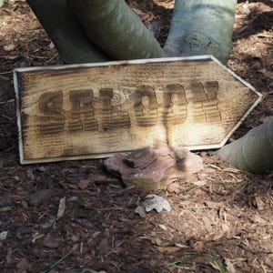 Könnte beinhalten: Ein Holzschild mit dem Wort "SALOON" darauf geschnitzt. Das Schild ist verwittert und hat einen rustikalen Look. Es liegt auf dem Boden mit einem Stein dahinter.