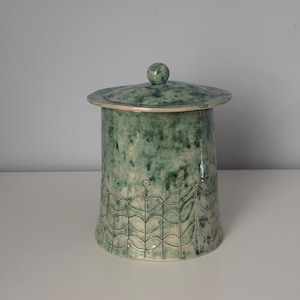 Può includere: Un contenitore cilindrico in ceramica con coperchio caratterizzato da una smaltatura maculata verde e bianca. La parte inferiore del contenitore è decorata con un motivo inciso ripetitivo di steli stilizzati e foglie circolari in verde scuro.
