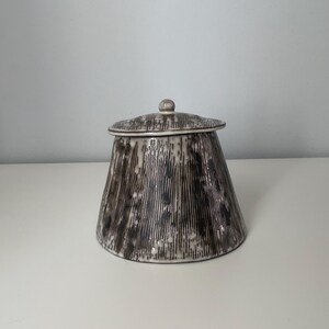 Peut inclure: Un pot en céramique avec un couvercle. Le pot est de couleur beige clair avec des lignes verticales noires et grises. Le couvercle a un petit bouton sur le dessus et un motif similaire au pot. Le pot est posé sur une surface blanche.