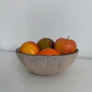 Peut inclure: Un bol beige avec un motif de lignes verticales texturées, rempli de fruits frais. Le bol contient un mélange d'oranges, de pommes et d'une mangue verte. Le bol de fruits est posé sur une surface blanche.