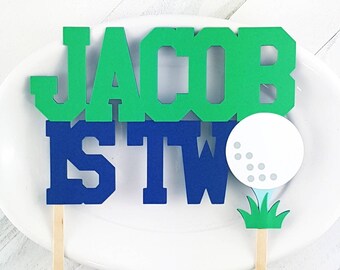 May include: Green and blue cake topper with the name "Jacob" and the words "is two" with a golf ball on top of a green stem.