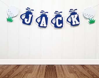 May include: A blue and white golf-themed banner spelling "JACK" with golf bag and golf ball cutouts. The banner is strung on a white string and is set against a white wall with a wooden floor.