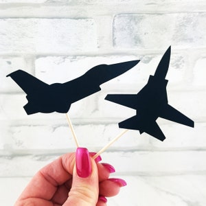 May include: Two black airplane-shaped cupcake toppers on wooden sticks. The background is a white brick wall. The airplane silhouettes are different designs. The toppers are ideal for decorating cakes.
