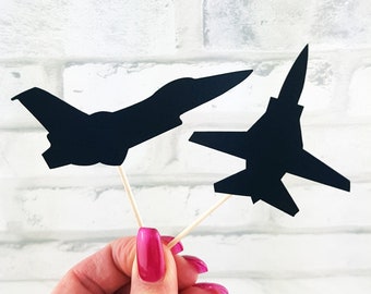 May include: Two black airplane-shaped cupcake toppers. The toppers are held by wooden sticks. The background is a white brick wall. The airplane silhouettes are different designs.