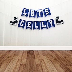 May include: A blue banner with white lettering that reads "LET'S CELLY" is strung across a white wall. Two black and white ice skates flank the banner. The banner is a festive decoration for a hockey-themed event.