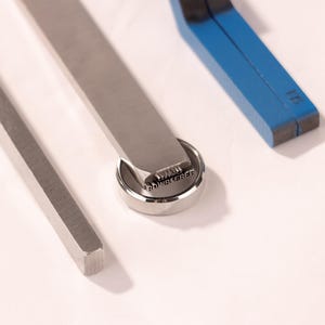 Op de afbeelding: Close-up van metalen gereedschappen, waaronder een stempel met de tekst "BOWREREE", een gereedschap met vierkante rand en een blauw gereedschap met het nummer "16". De gereedschappen zijn gerangschikt op een witte ondergrond, wat hun metalen afwerking en precisie benadrukt.