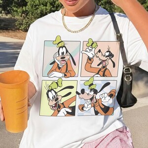 Puede incluir: Una camiseta blanca con un estampado gr&aacute;fico de cuatro paneles distintos, cada uno mostrando al personaje de dibujos animados Goofy en diferentes poses, vistiendo un su&eacute;ter naranja, chaleco negro y un sombrero verde sobre fondos pastel.