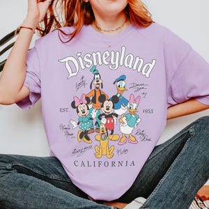 以下が含まれることがあります： ディズニーランドのロゴと、ミッキーマウス、ミニーマウス、ドナルドダックなどのディズニーキャラクターのサインが特徴のライラック色のTシャツ。「CALIFORNIA」と「1955」の文字も含まれています。