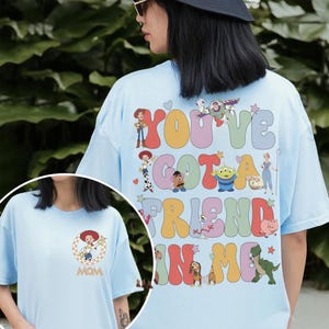 Peut inclure: Un t-shirt bleu clair avec un grand motif au dos portant le texte "YOU'VE GOT A FRIEND IN ME" en lettres ondul&eacute;es color&eacute;es entour&eacute;es de personnages de Toy Story. La poitrine affiche un graphique circulaire de Jessie et le mot "MOM".