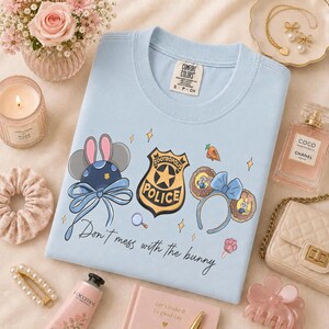 May include: A light blue t-shirt featuring a graphic print of a Zootropolis police badge, two pairs of mouse ears, a carrot, and the text "Don't mess with the bunny" in black script. The shirt has a crew neckline and a Comfort Colors brand label.